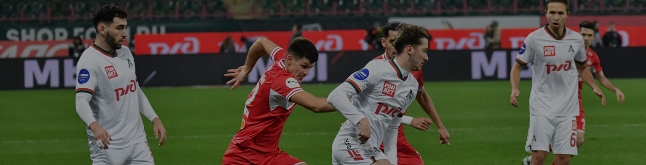 На изображении несколько футболистов в белой и красной форме играют в футбол на поле. Один игрок в белой форме с мячом, против него активно защищаются. На заднем плане видны зрители и баннеры